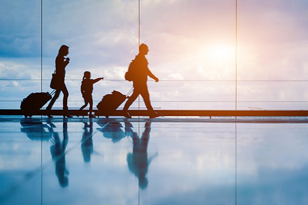 Family at airport travelling with young child and luggage walking to departure gate, girl pointing at airplanes through window, silhouette of people, abstract international air travel concept